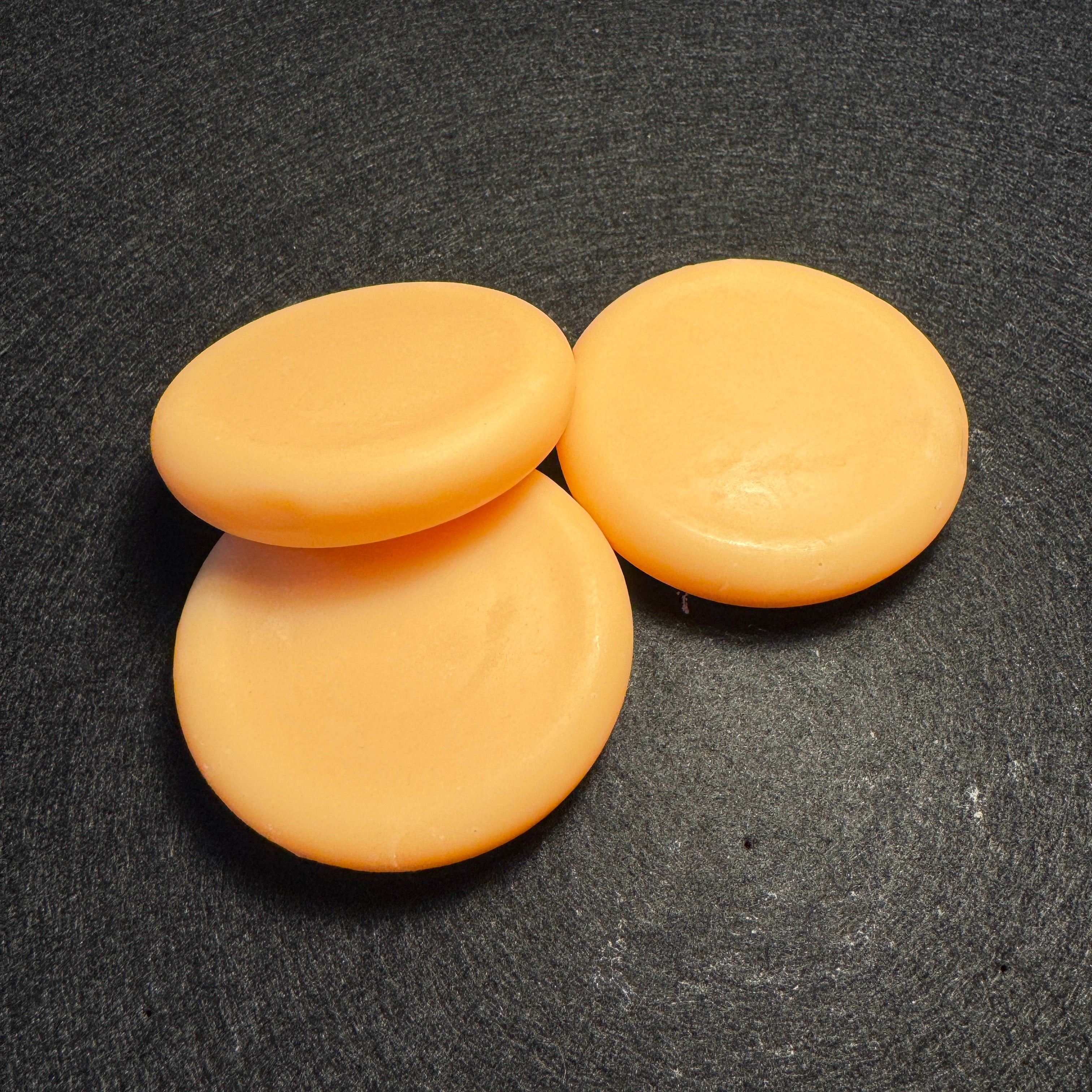 Three round, pale peach-colored soap bars arranged on a dark textured surface.