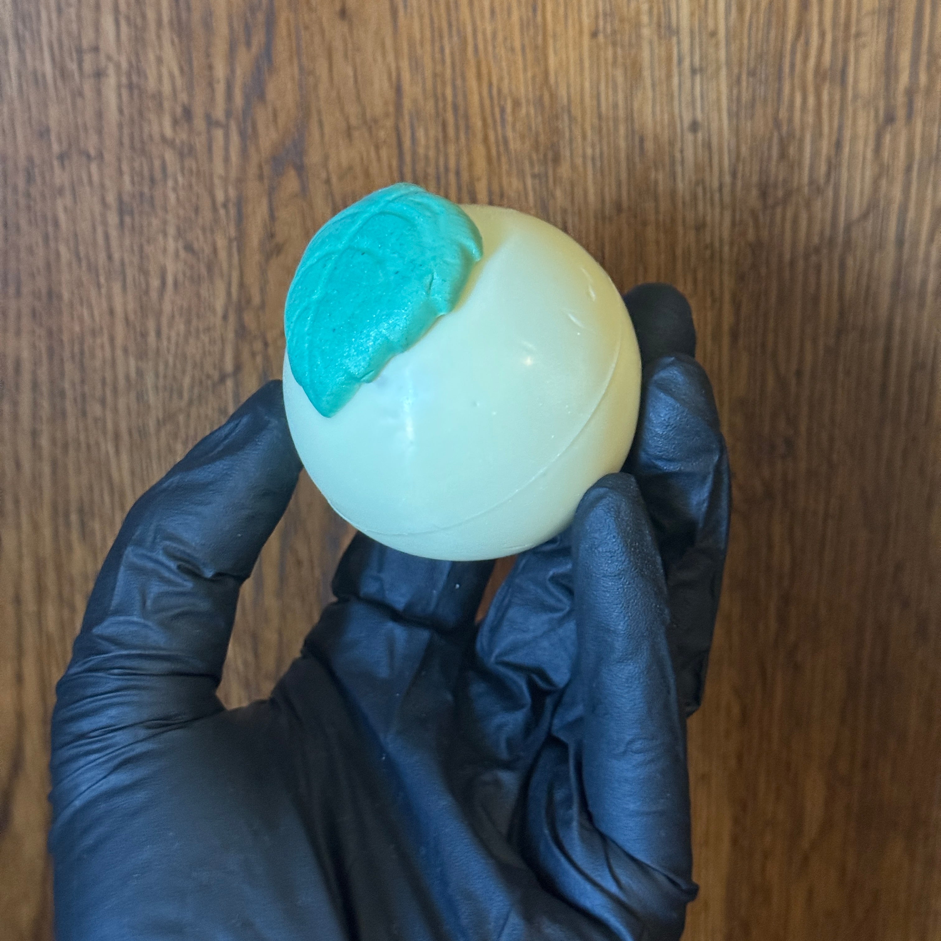 Hand wearing a black glove holding a light yellow balls of soap topped with a green soap leaf.