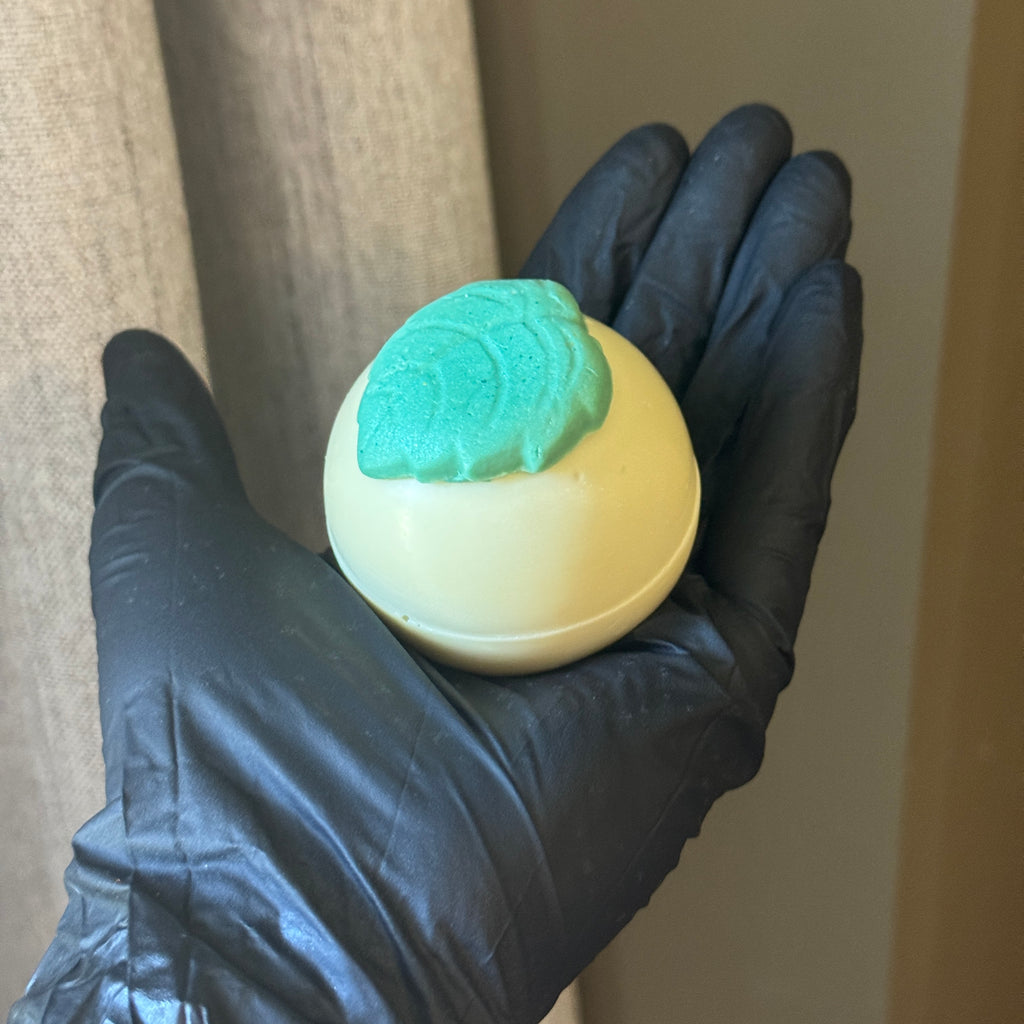 Hand wearing a black glove holding a light yellow balls of soap topped with a green soap leaf.
