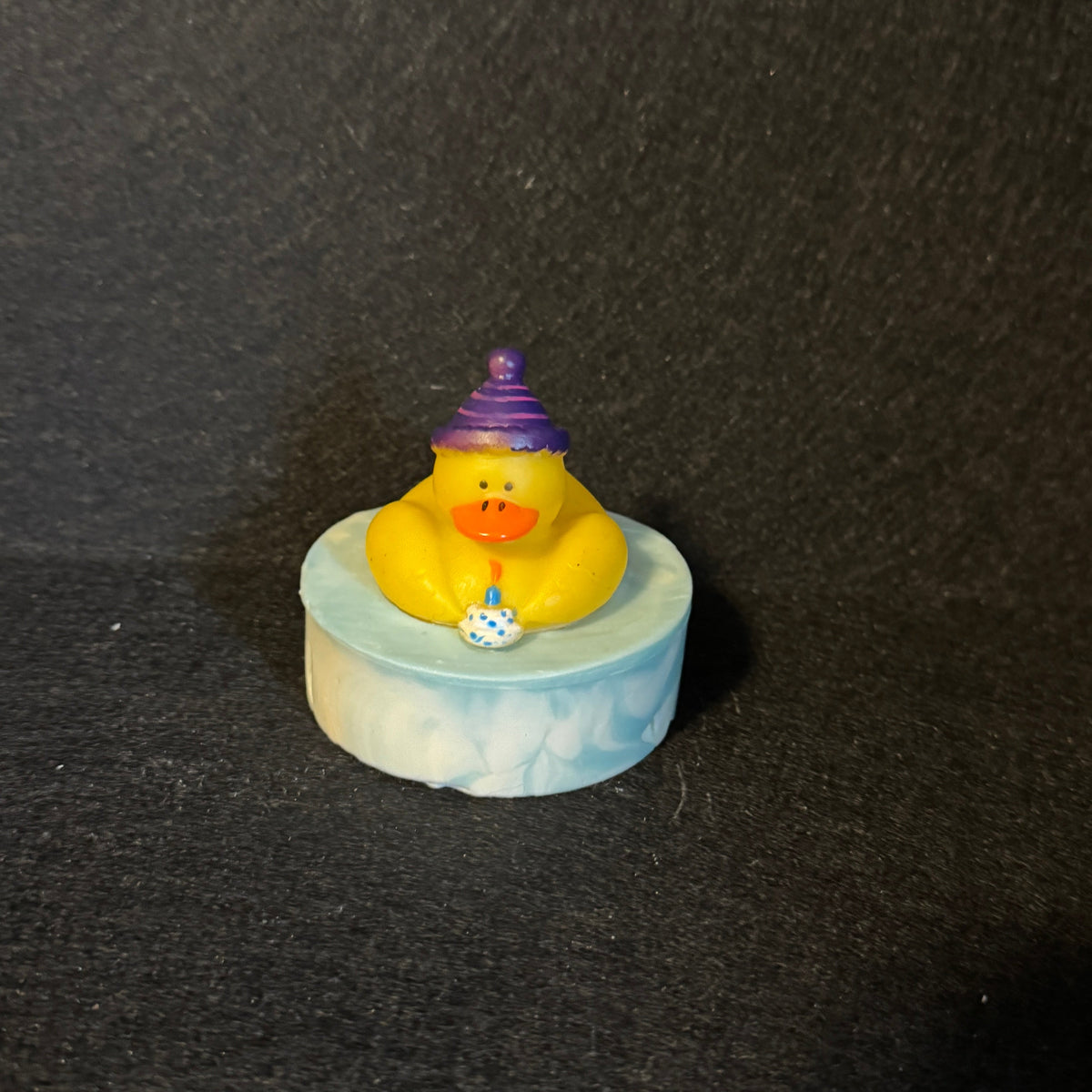 A small yellow rubber duck wearing a purple party hat, sitting on a round light blue and white marbled base against a dark textured background.