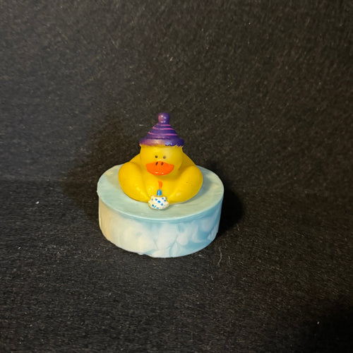 A small yellow rubber duck wearing a purple party hat, sitting on a round light blue and white marbled base against a dark textured background.