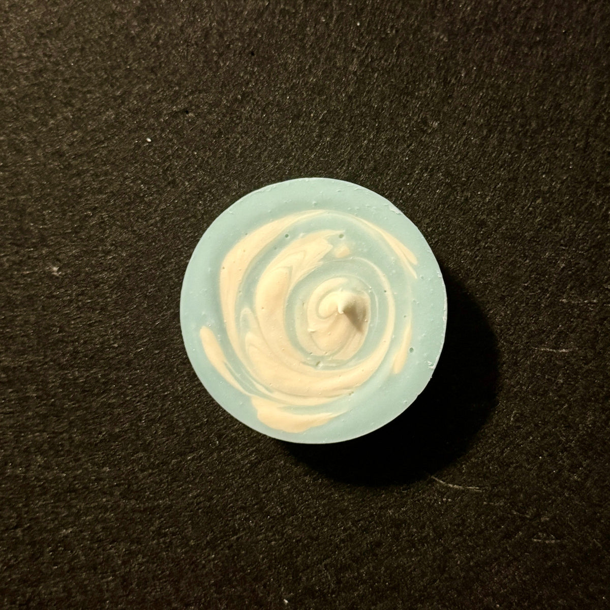 A small round translucent light blue item with a white spiral pattern in the center, placed on a dark textured background.