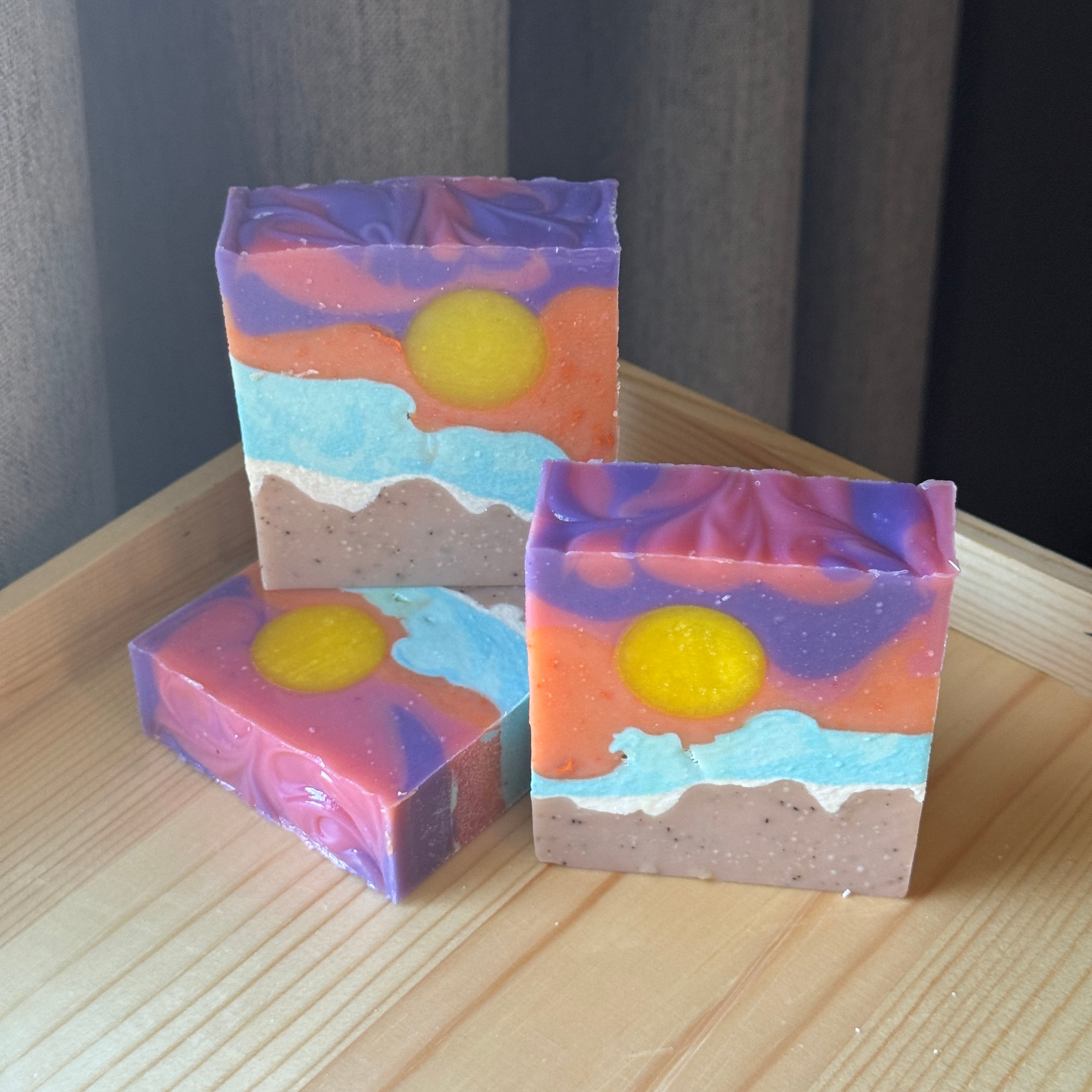 Three colorful soap bars with sun and water and beach designs on a wooden surface.