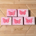 Five pink square shaped soaps with a butterfly embed in a grid on a wooden surface