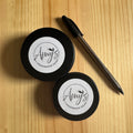 Two containers of 'Amy's Handmade Soap' on a wooden surface with a pen.
