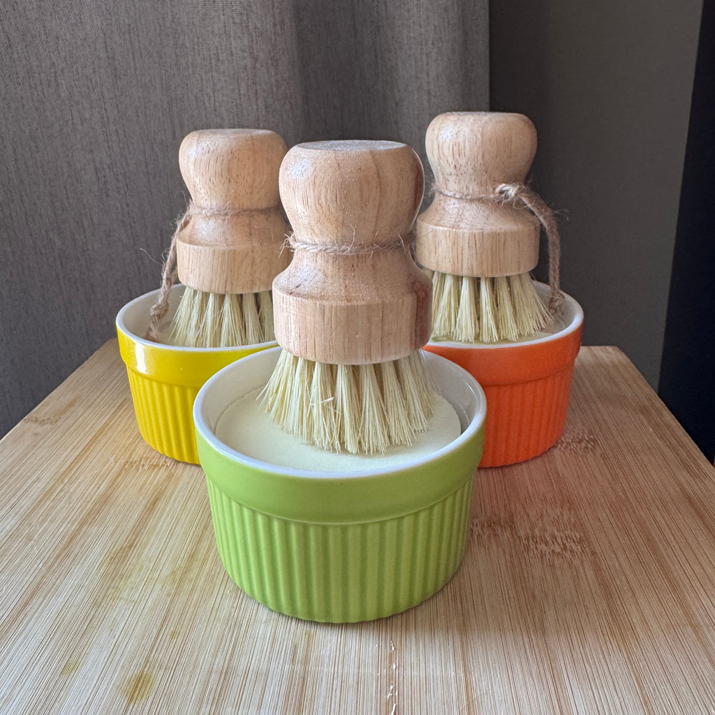 Set of three wooden-handled dish brushes in colourful ramekin on a wooden surface.