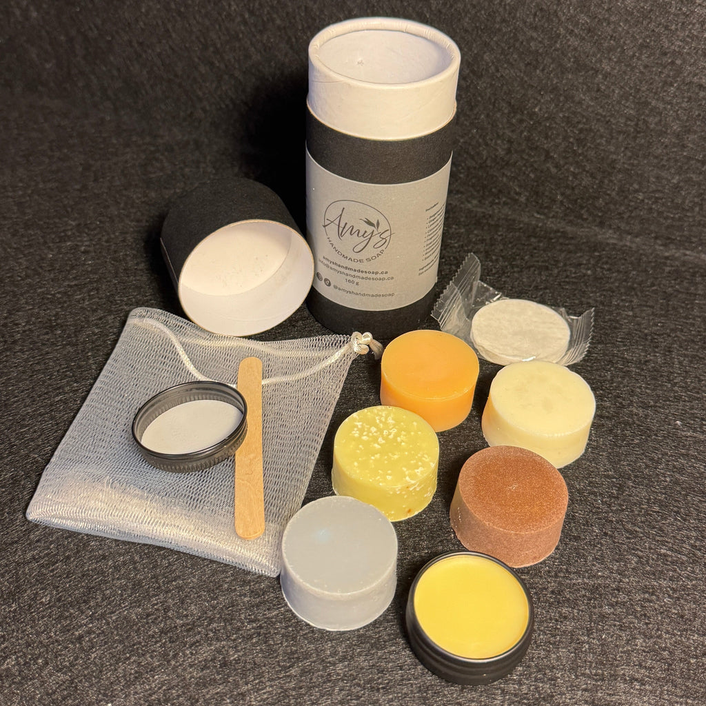 A set of small cosmetic or skincare jars and containers arranged on a dark textured surface. The set includes a tall white cylindrical bottle with a dark gray band, several small round jars in white, yellow, orange, and brown, a small clear pouch with a metal ring and wooden spatula, and a small metal pot with white contents.