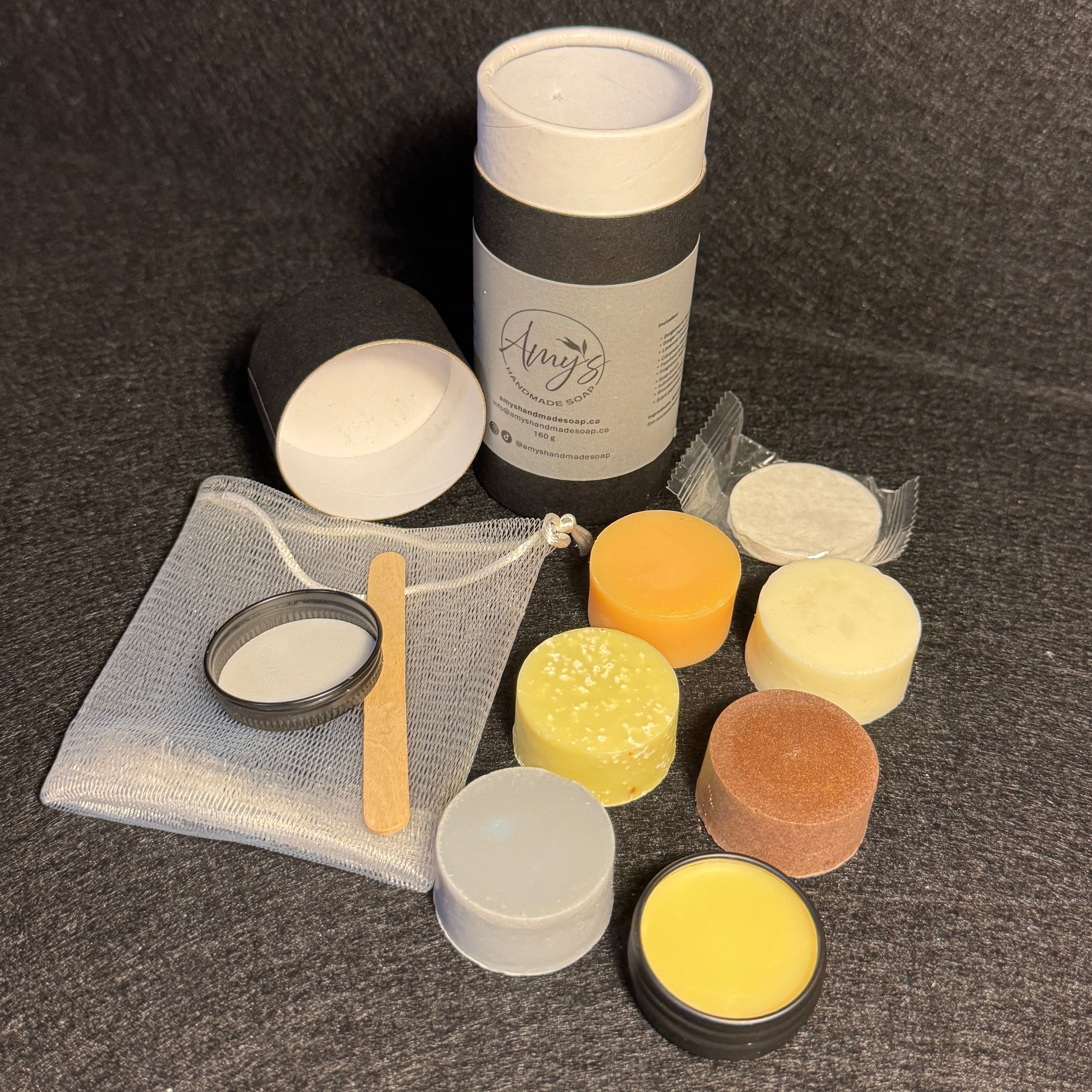 A set of small cosmetic or skincare jars and containers arranged on a dark textured surface. The set includes a tall white cylindrical bottle with a dark gray band, several small round jars in white, yellow, orange, and brown, a small clear pouch with a metal ring and wooden spatula, and a small metal pot with white contents.