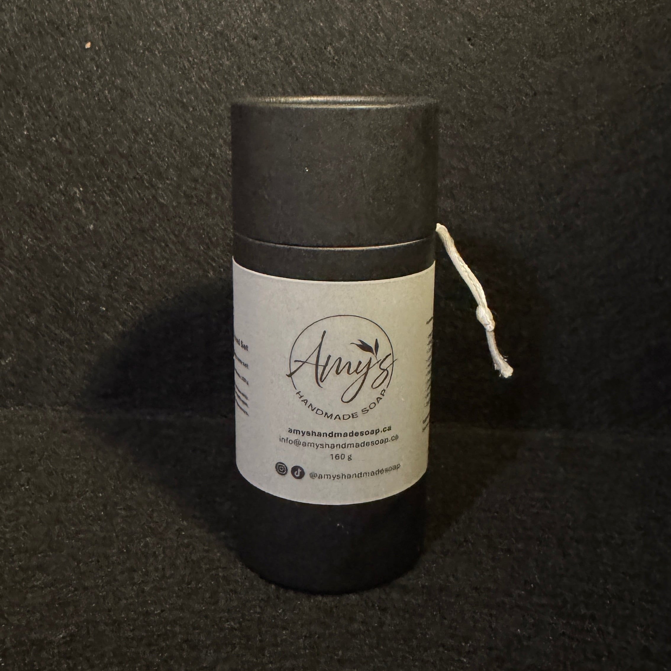 A black cylindrical candle jar with a white paper label reading 'Amy's Handmade Soap' and a white wick protruding from the side, set against a dark textured background.