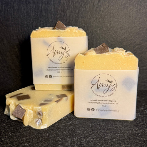 Three bars of handmade soap by Amy's Handmade Soap, featuring a pale yellow creamy base with embedded dark chocolate chunks, displayed on a dark textured background.