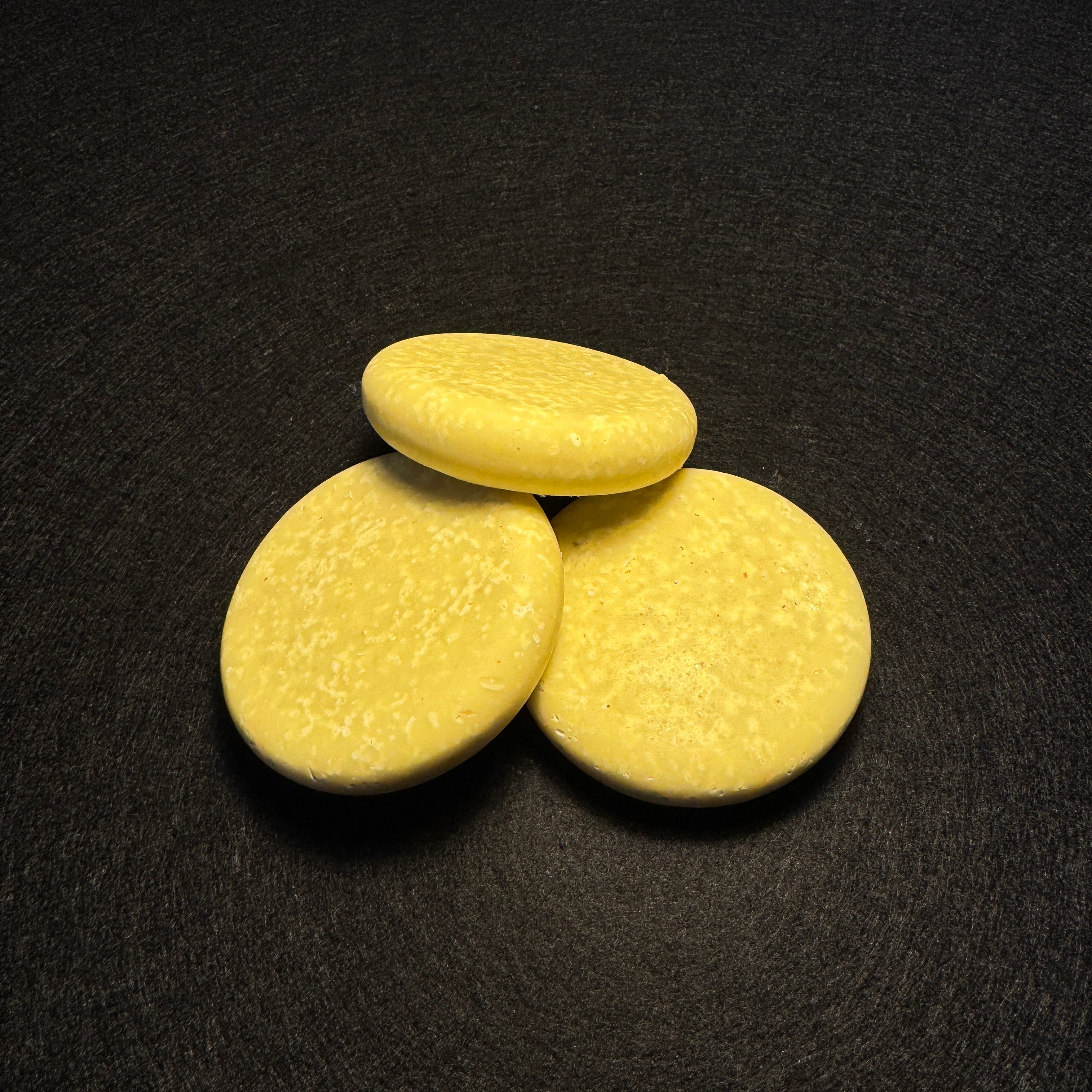 Three round, pale yellow soap bars arranged in a small pyramid on a dark textured background.