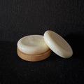 Two oval cream-colored soap bars with a light wood base, displayed on a dark textured background.