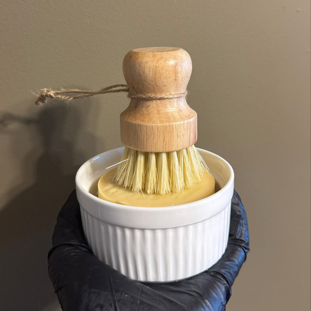 Hand holding a white ramekin with a wooden brush and yellow soap against a plain background