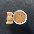 A small cleaning brush with a light wood handle and cream bristles placed beside a round white container with a bamboo lid, photographed on a dark textured surface.