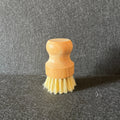 A small wooden-handled cleaning brush with pale yellow bristles, standing upright on a dark textured surface against a dark gray backdrop.