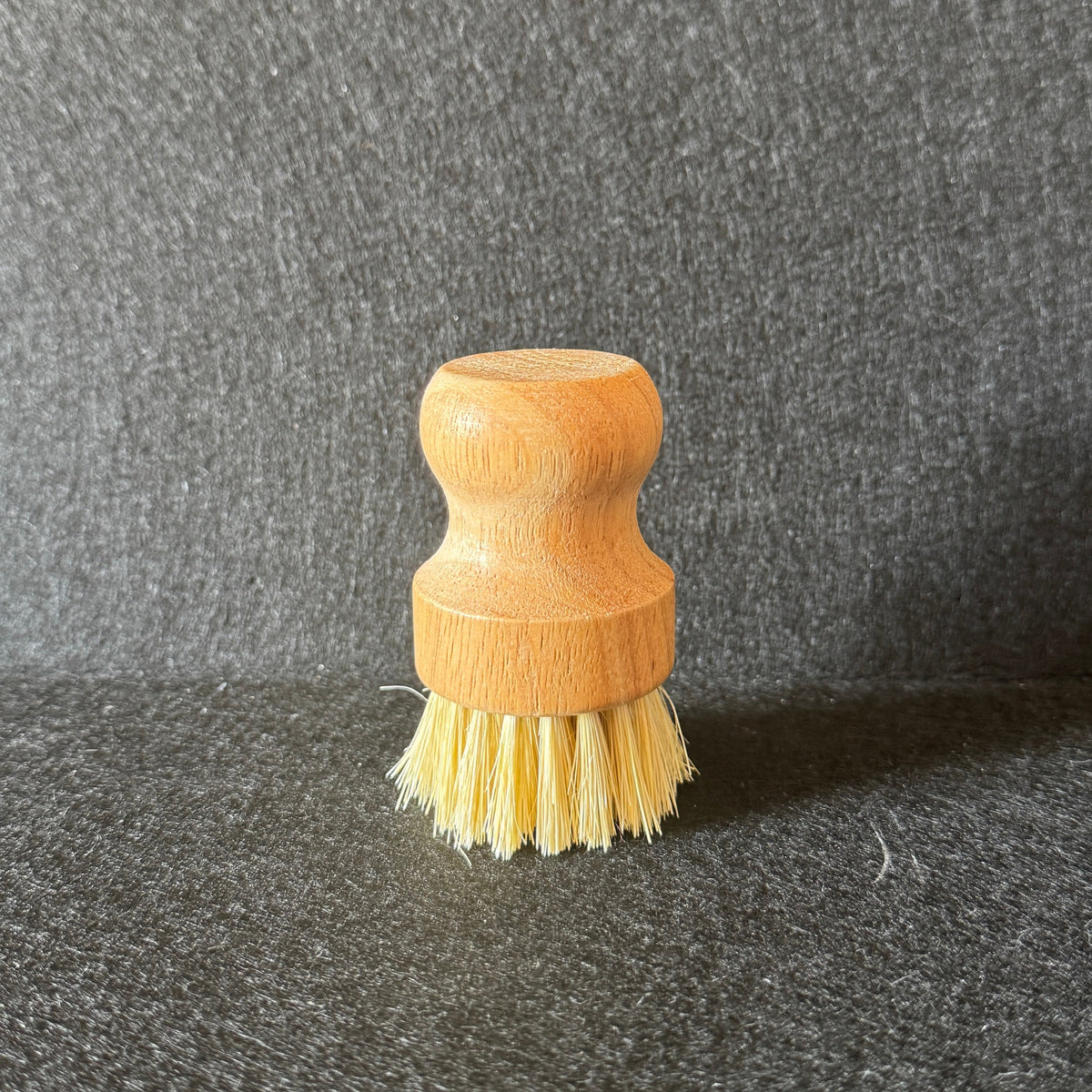 A small wooden-handled cleaning brush with pale yellow bristles, standing upright on a dark textured surface against a dark gray backdrop.