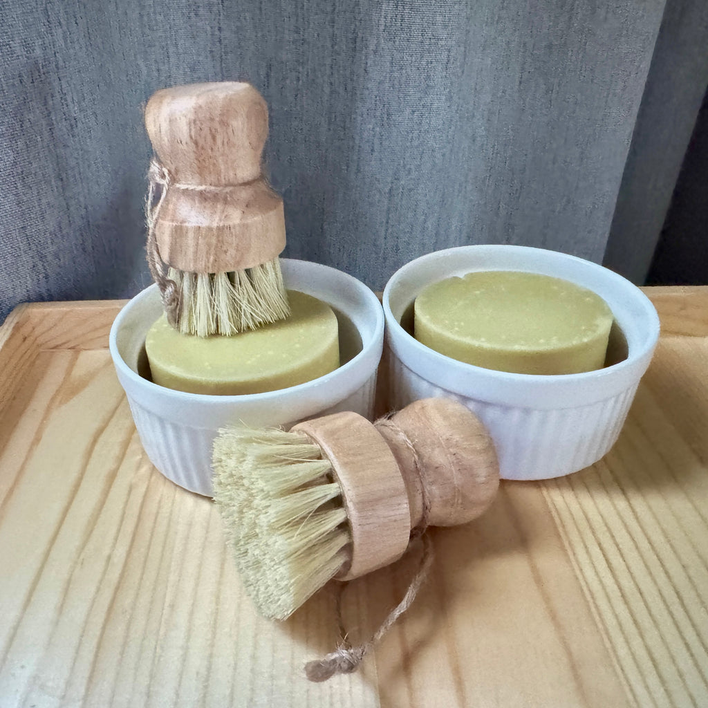 Two wooden brushes and two bars of soap in white containers on a wooden surface.