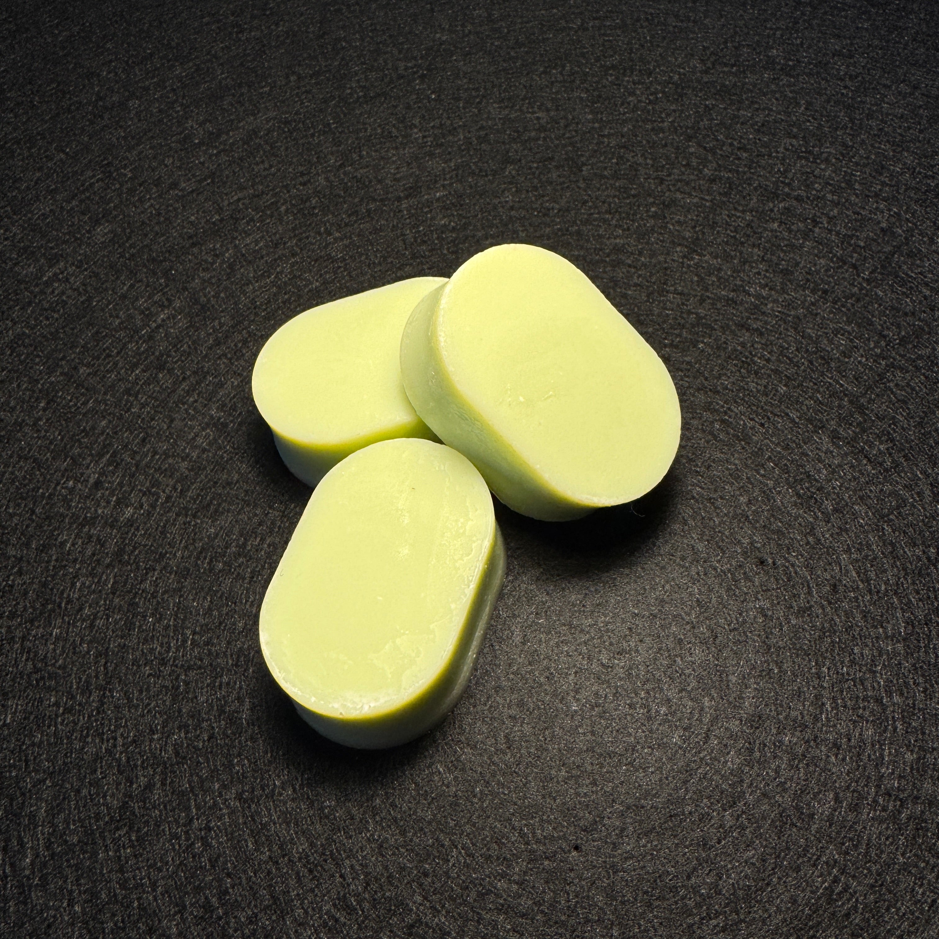 Three pale green oval-shaped soap bars arranged on a dark textured surface.