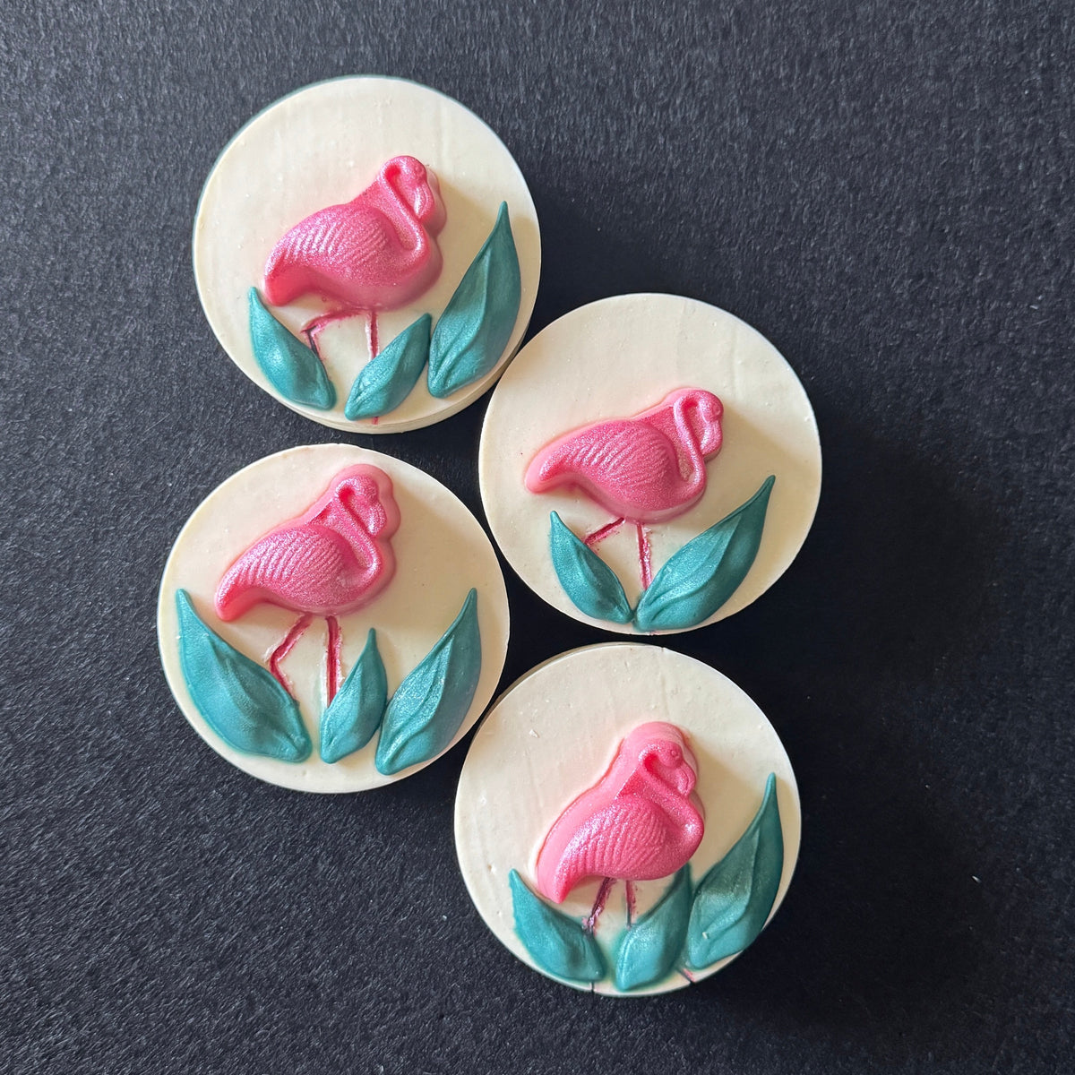Four round white soaps featuring a pink flamingo and teal leaves design, arranged on a dark textured background.
