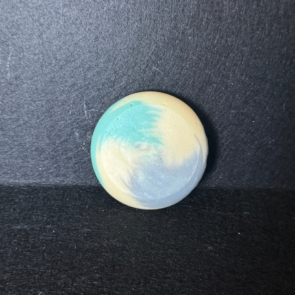A small round soap with a marbled swirl pattern of light blue, cream, and white, resting on a dark textured surface.