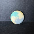 A small round soap with a marbled swirl pattern of light blue, cream, and white, resting on a dark textured surface.