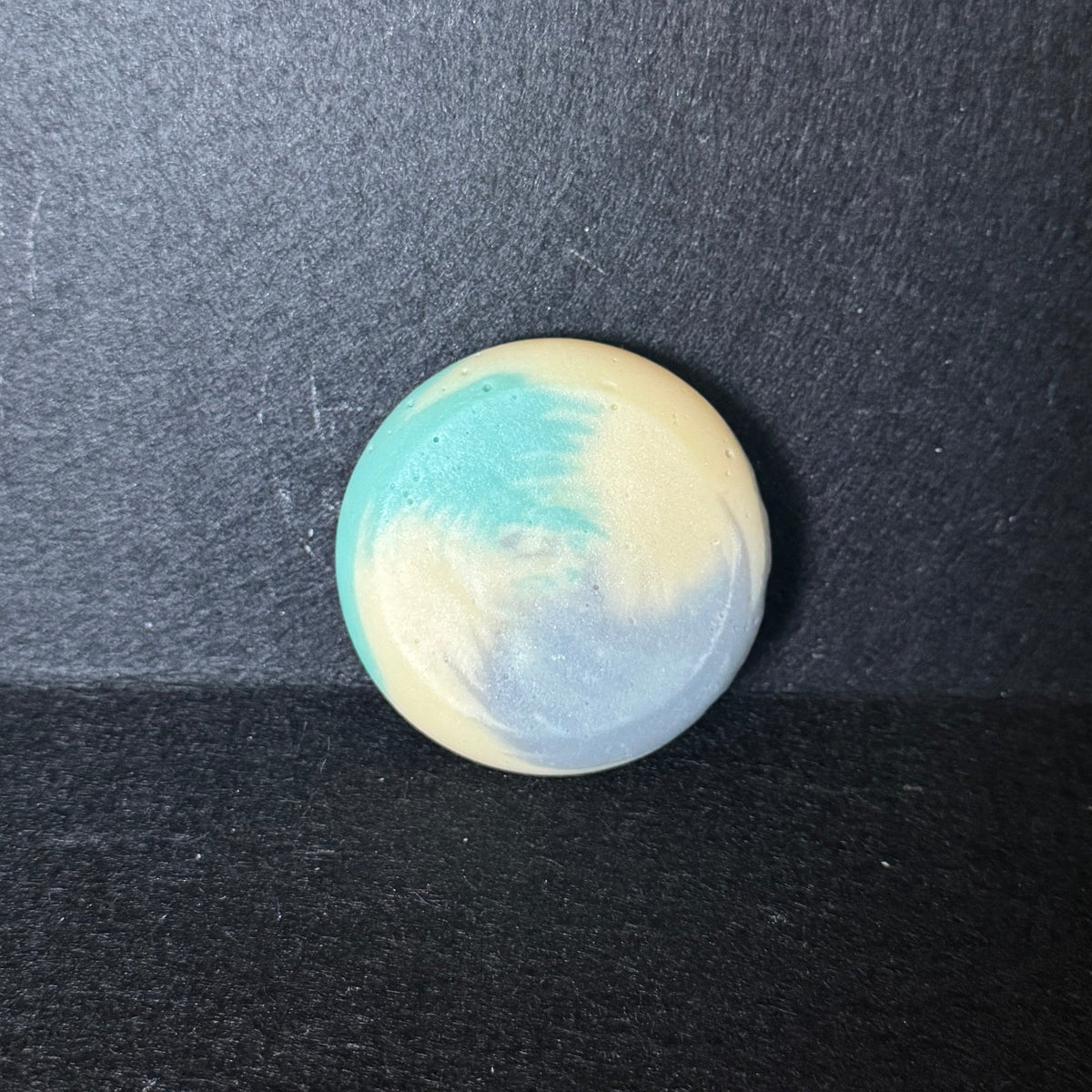 A small round soap with a marbled swirl pattern of light blue, cream, and white, resting on a dark textured surface.