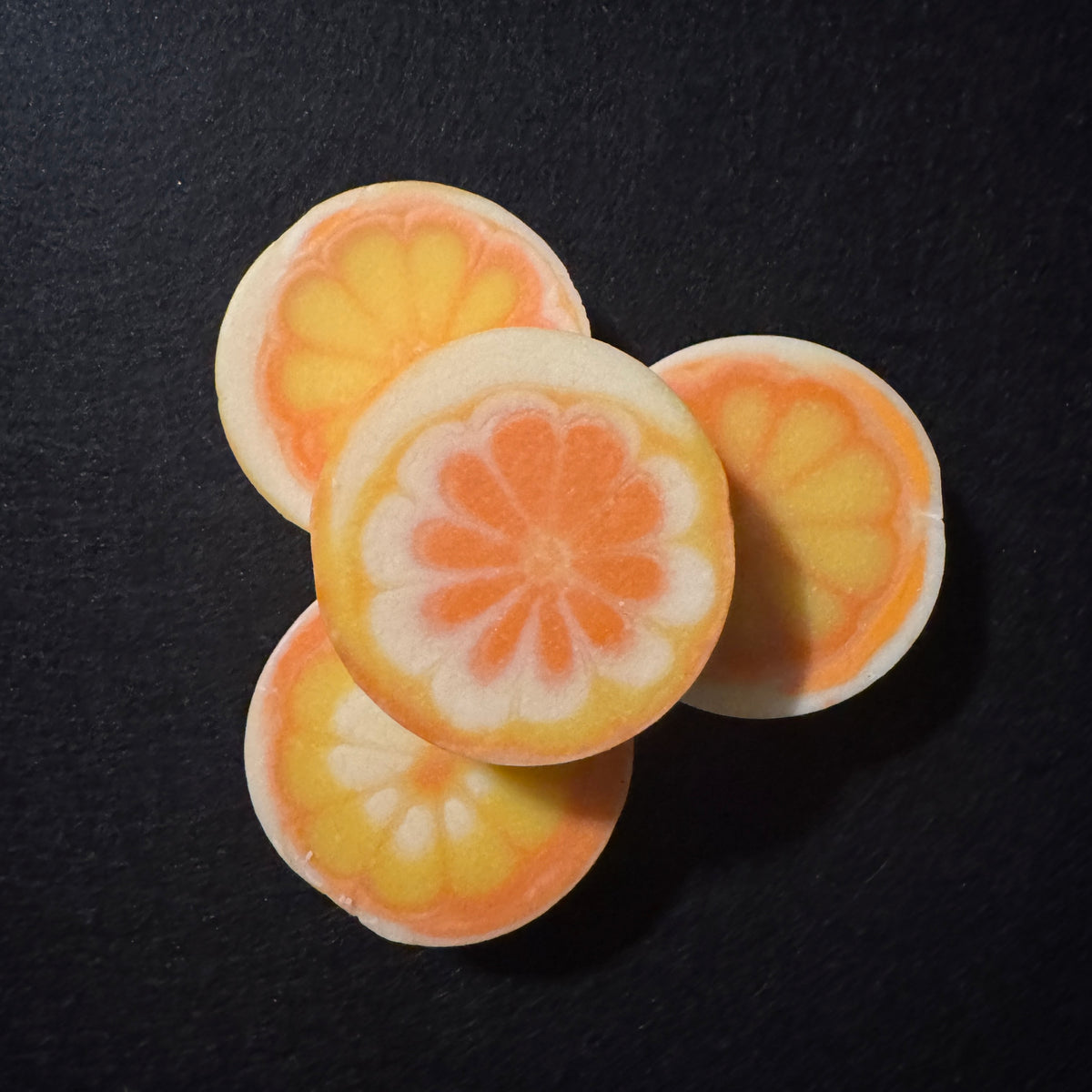 Four round, sliced soaps with a layered yellow, orange, and white citrus sliced pattern, arranged on a dark black background.