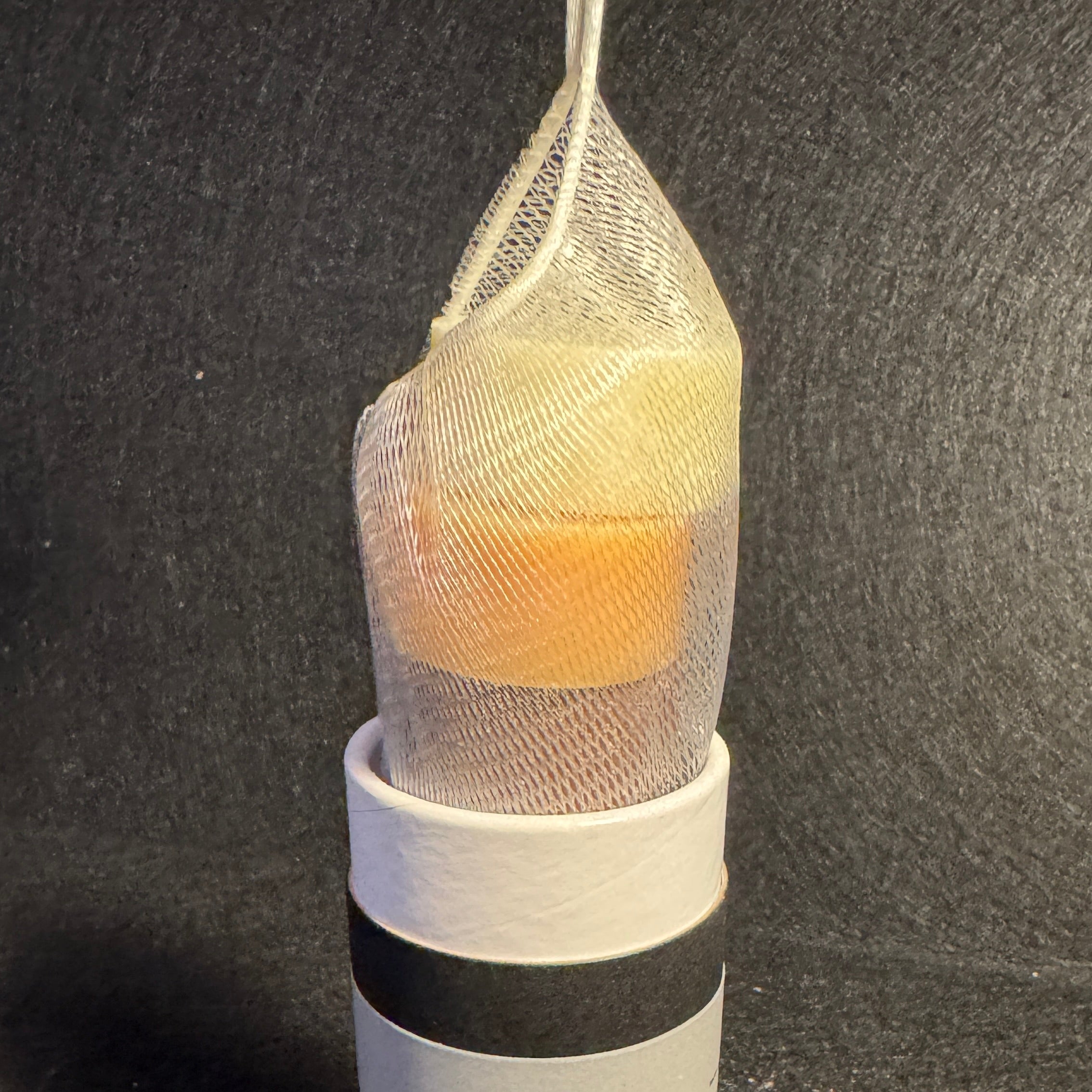 A bath product in a white and black cylindrical container with a mesh bag containing a yellow-orange-brown-white soaps, photographed on a dark textured surface.