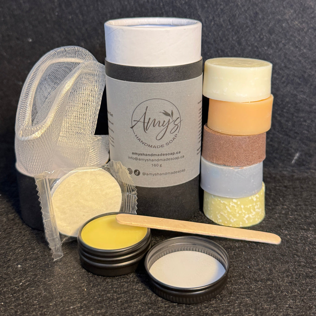 A handmade soap kit displayed on a dark textured background, including a cylindrical tin labeled 'Amy's', stacked colored soaps, a small metal tin with yellow soap, an open tin with white soap, a wooden spatula, and a mesh soap bag.