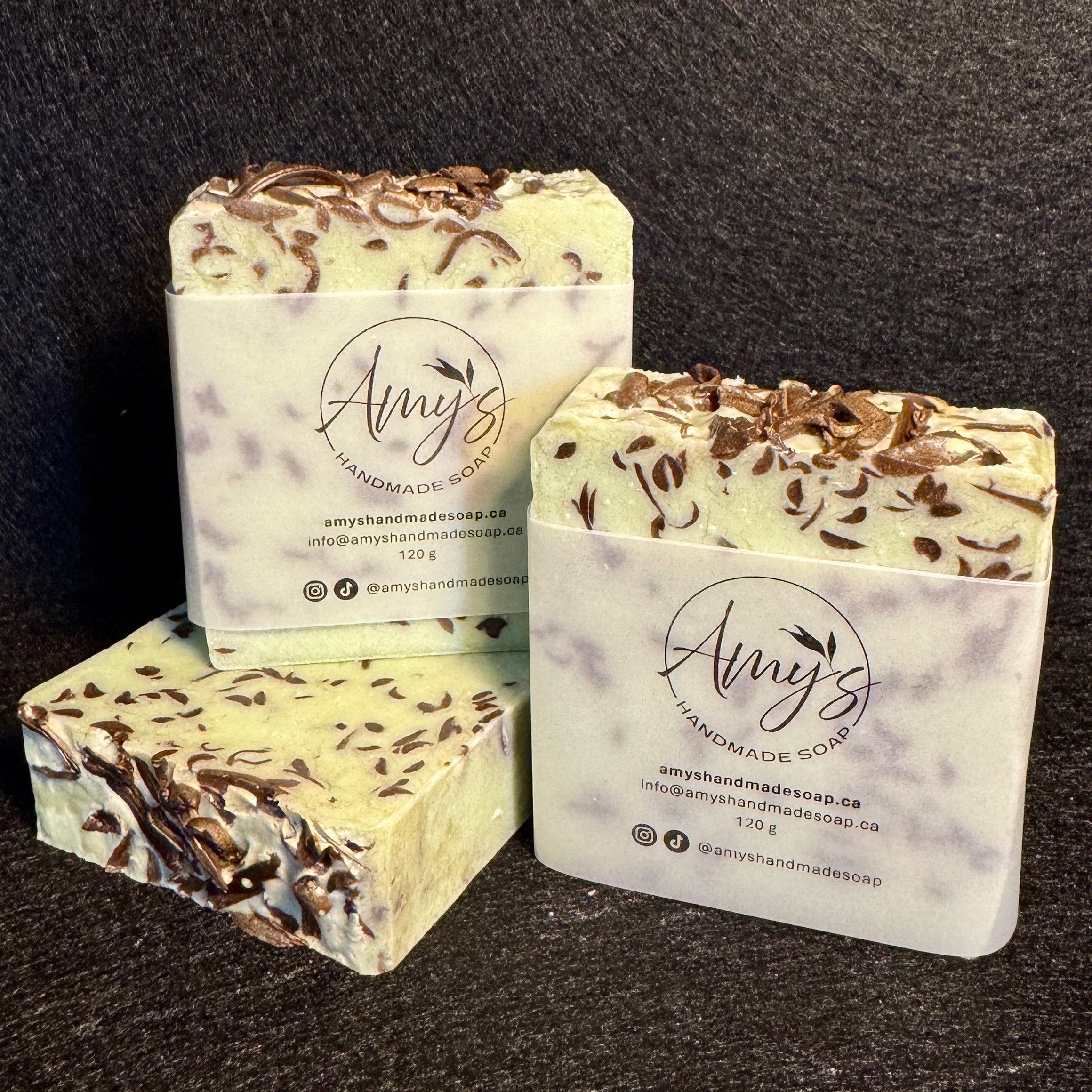 Three handmade soap bars wrapped in white paper labels reading Amy's Handmade Soap, arranged on a dark textured background. The soaps are pale green with chocolate shavings and topped with dark chocolate shavings.