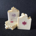 Three bars of handmade soap in off-white with a red maple leaf design, displayed on a dark textured background. One bar is standing upright with its label visible, which reads 'Amy's Handmade Soap Co'.