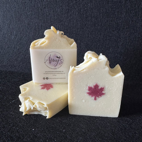 Three bars of handmade soap in off-white with a red maple leaf design, displayed on a dark textured background. One bar is standing upright with its label visible, which reads 'Amy's Handmade Soap Co'.