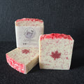 Three handmade soap bars displayed on a dark textured background. One bar is standing upright with a white paper label that reads 'Amy's Handmade Soap', while two other bars are placed in front, showing the creamy off-white soap base speckled with red and pink, and one with a red maple leaf design on top.