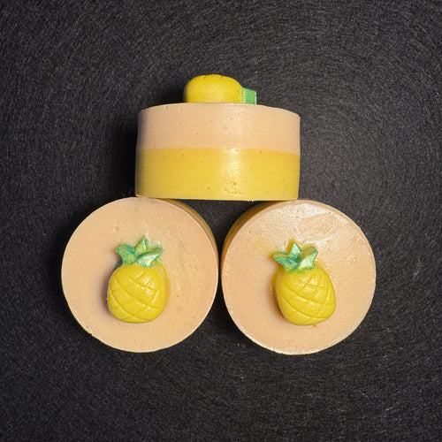 Three pineapple-themed soap bars arranged on a dark textured background, with one upright bar and two flat circular slices, each featuring a yellow pineapple detail on a beige base.