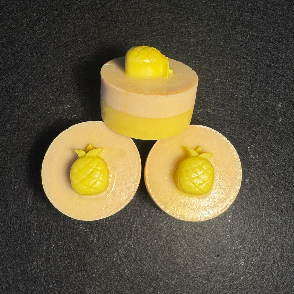 Three small round yellow and cream-colored soaps arranged on a dark gray surface, with one standing upright in the back and two lying flat in front. Each item has a small pineapple-shaped detail on top.