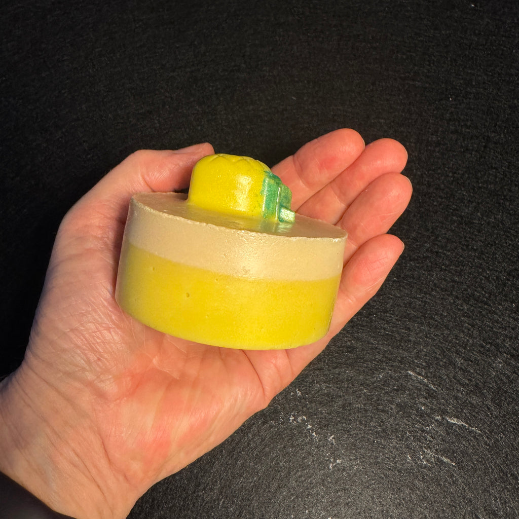 A hand holding a small round yellow and cream-colored soap bar with a pinapple-shaped topper, set against a dark textured background.