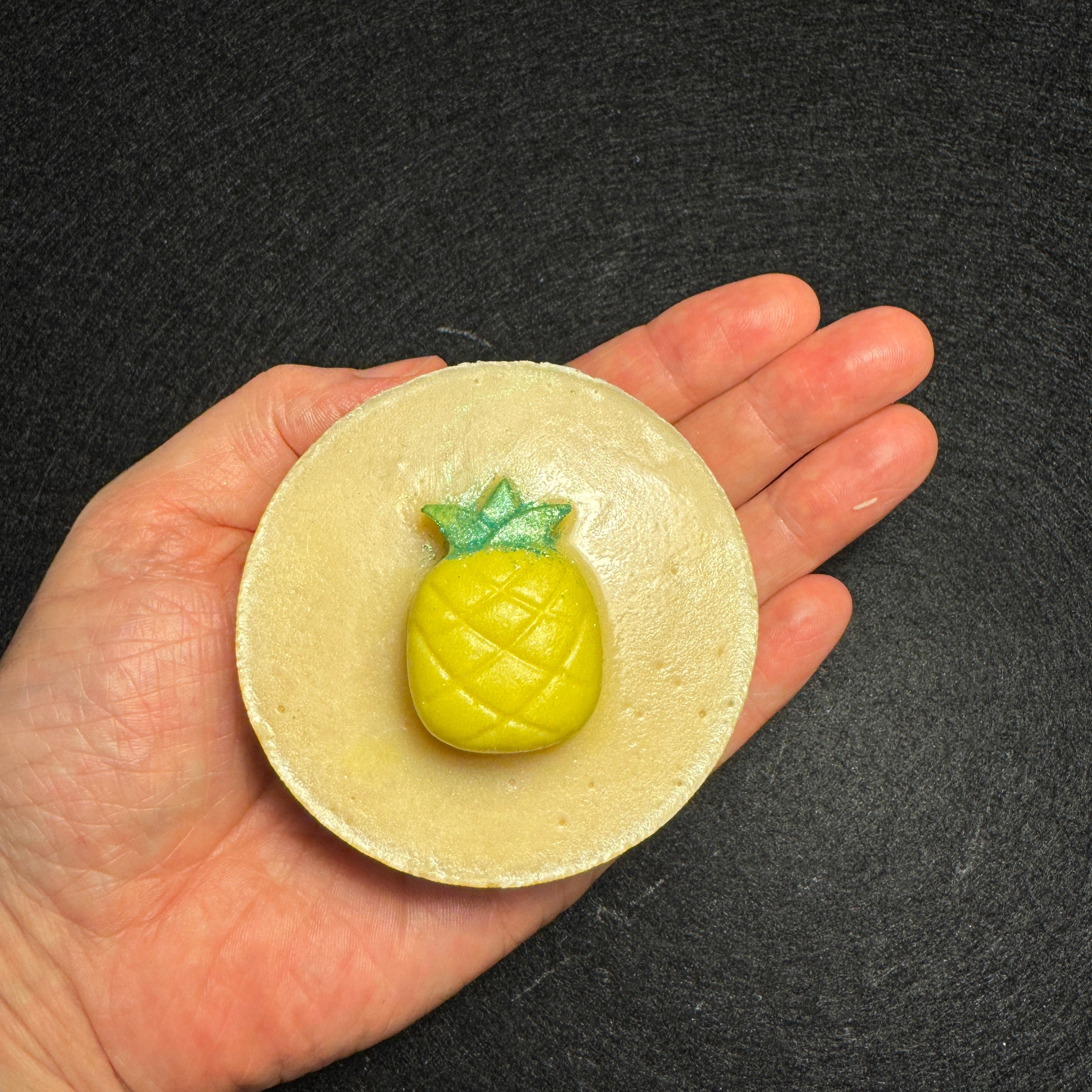 A hand holding a round, pale yellow soap to show its size, featuring a small, detailed yellow pineapple topper in the center with green leaves.