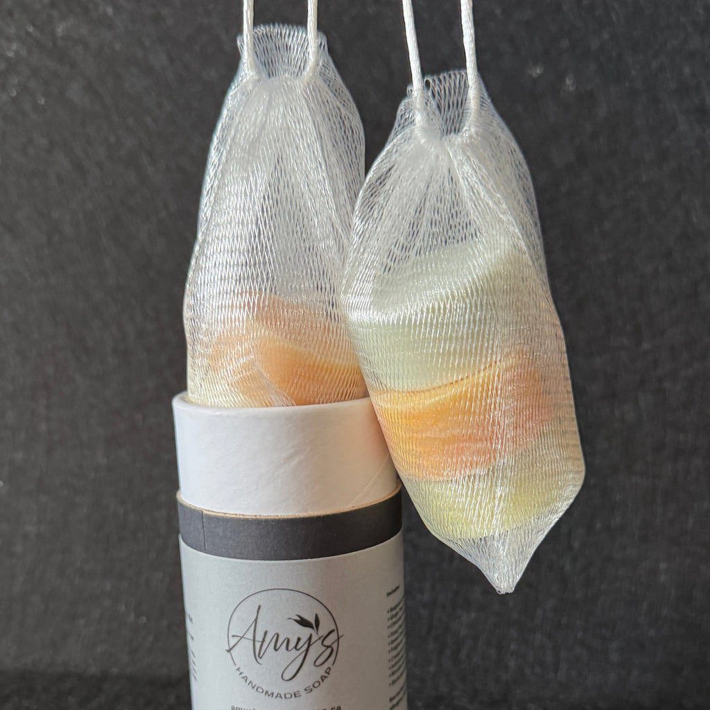 Two mesh bath soak bags filled with orange and yellow soaps, hanging from white cords above a cylindrical white and gray container labeled Amy's Handmade Soap on a dark textured background.