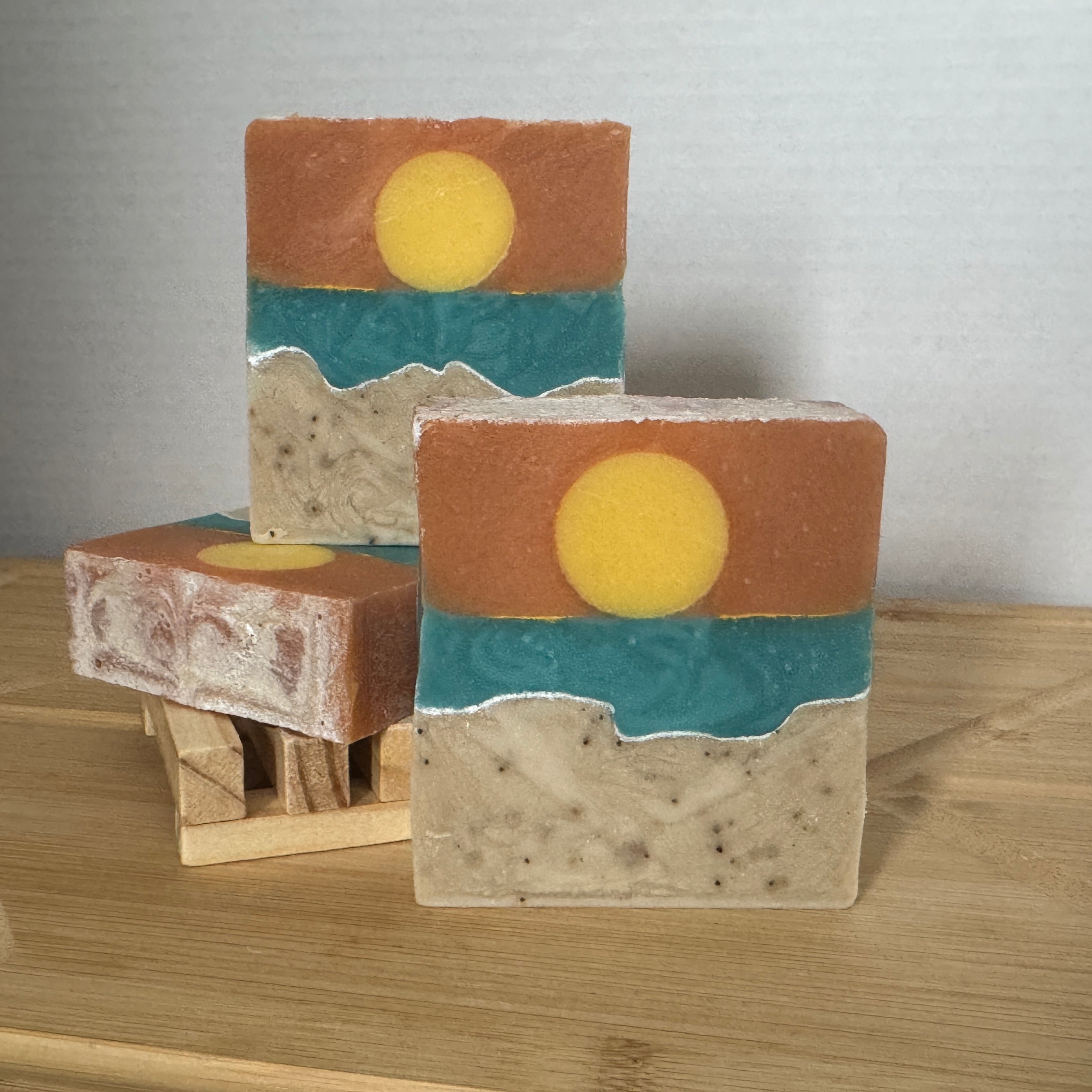Three soap bars with artistic layered designs resembling a sea landscape on a wooden surface.