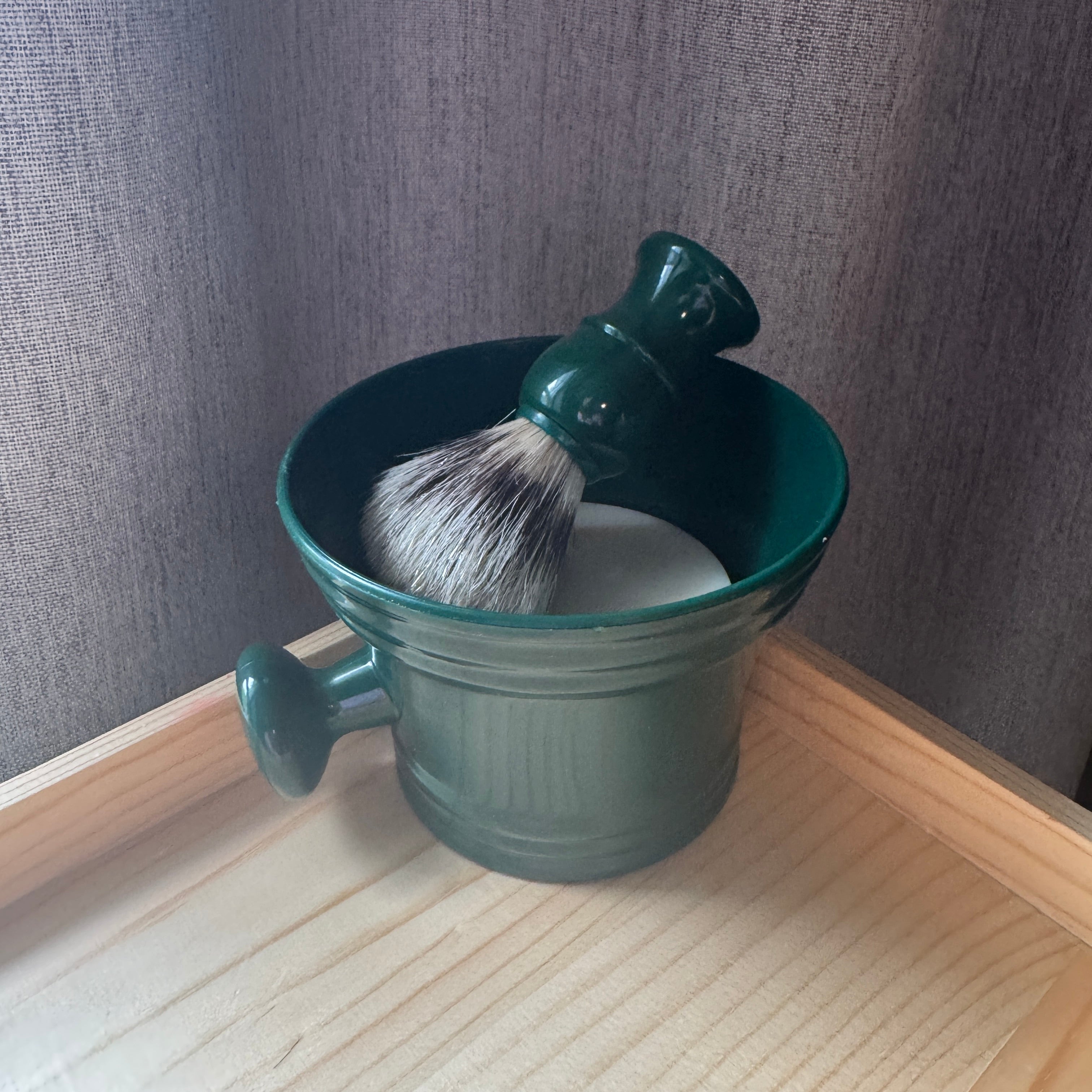 Green shaving mug with a brush and lather on a wooden surface against a gray wall.