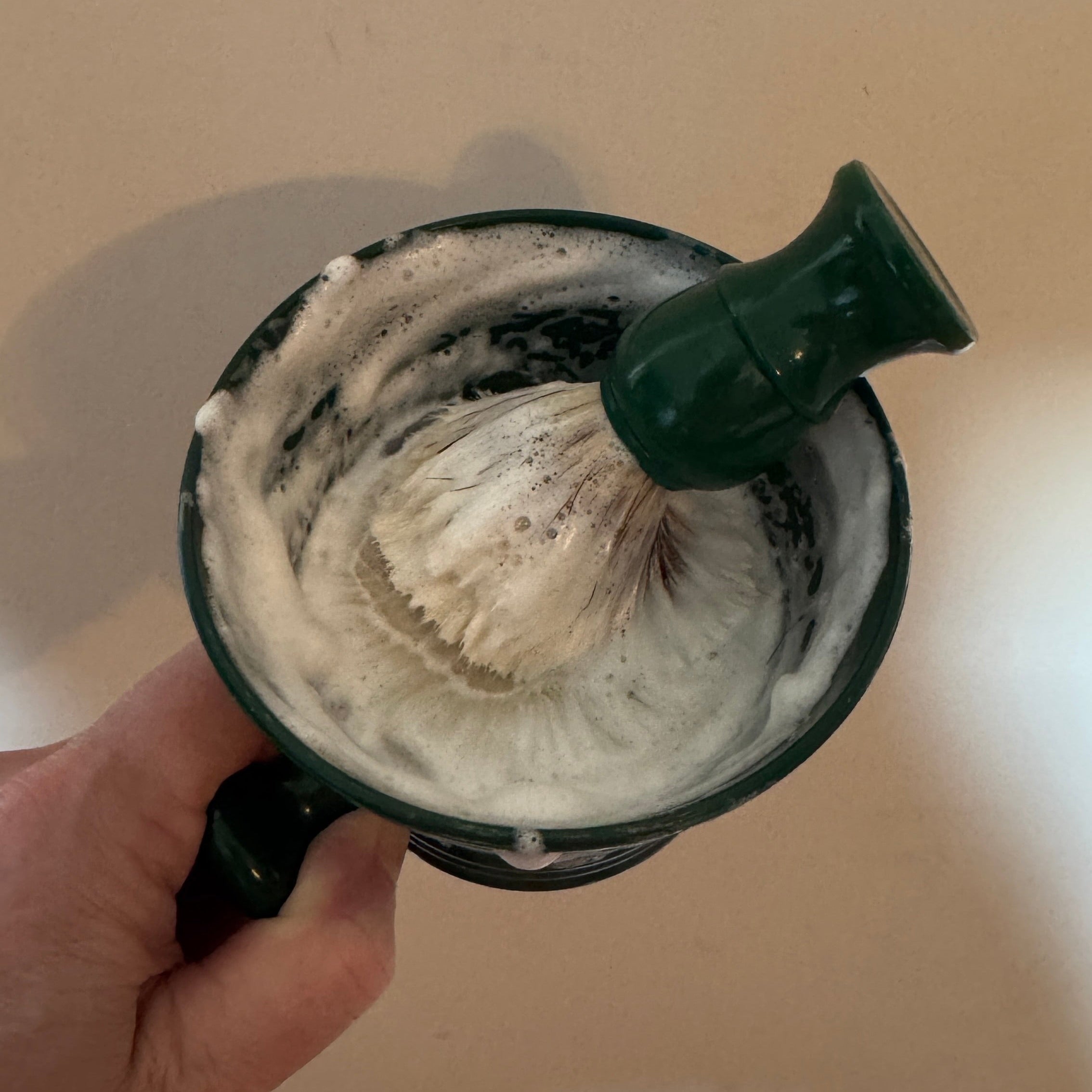 Person holding a shaving brush with lather in a mug against a neutral background