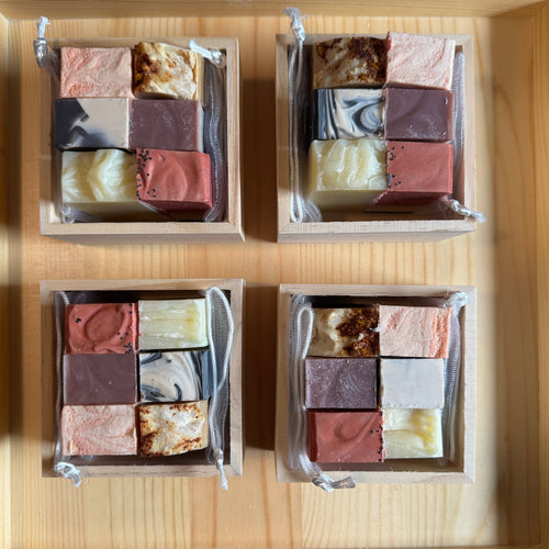 Four wooden boxes containing square pieces of soap arranged in a grid pattern on a wooden surface.