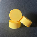 Three small yellow cylindrical soaps arranged in a stack on a dark textured surface with a bright light patch in the upper left.