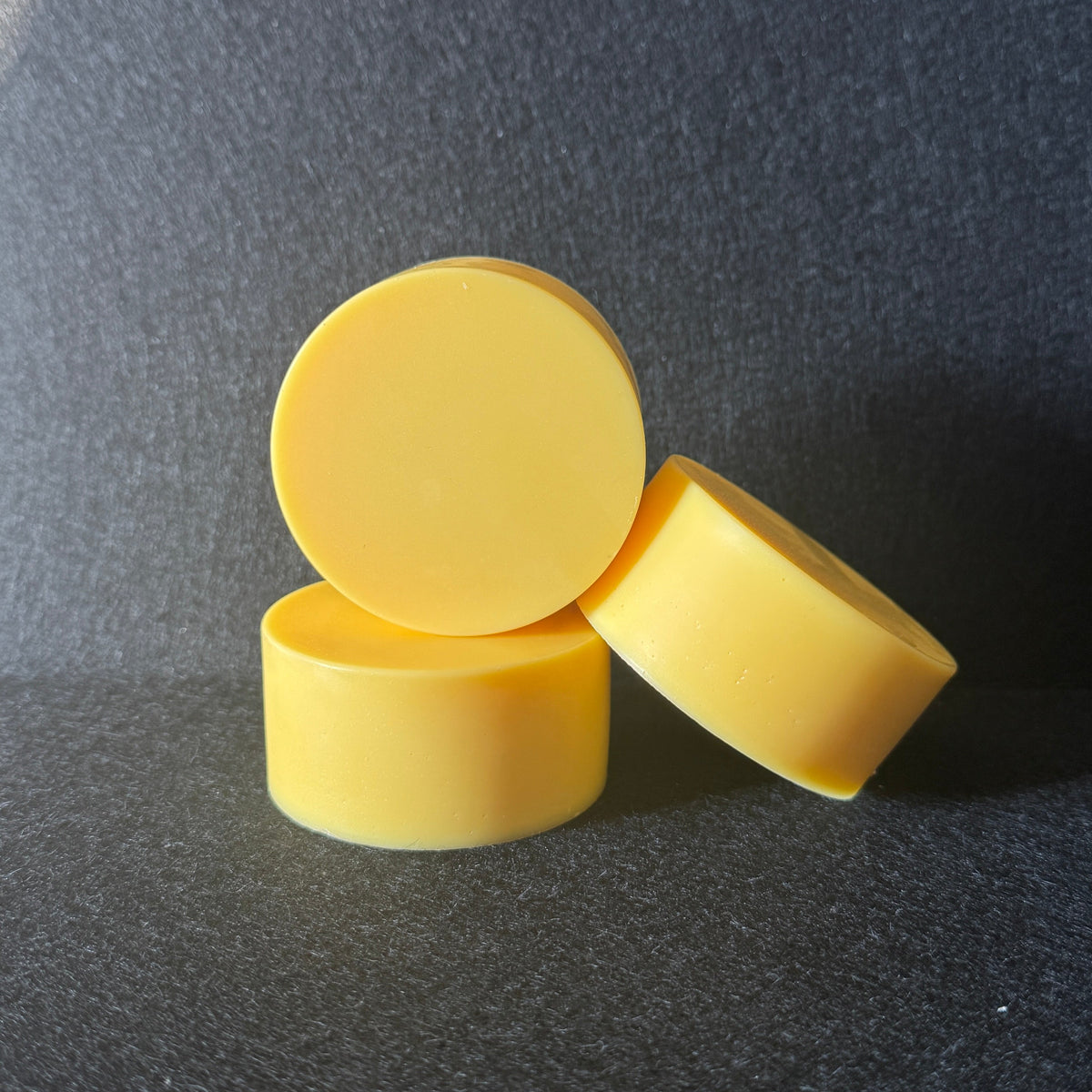 Three small yellow cylindrical soaps arranged in a stack on a dark textured surface with a bright light patch in the upper left.