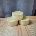 Three cylindrical beige soap bars stacked on a wooden surface with a gray textured background
