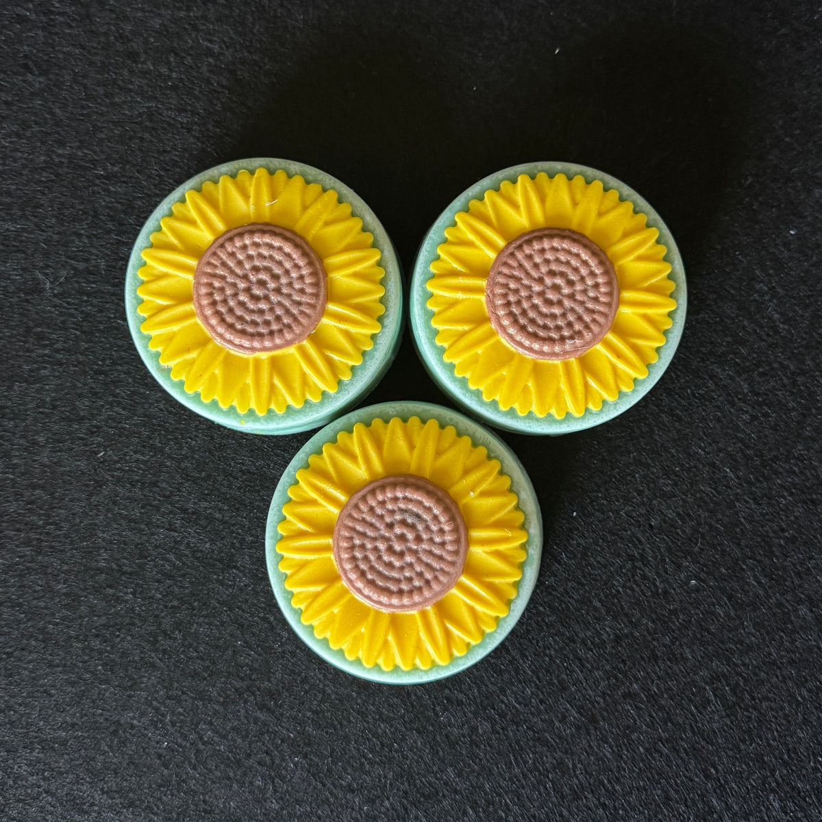 Three circular sunflower-shaped soaps arranged in a triangle on a dark textured background.