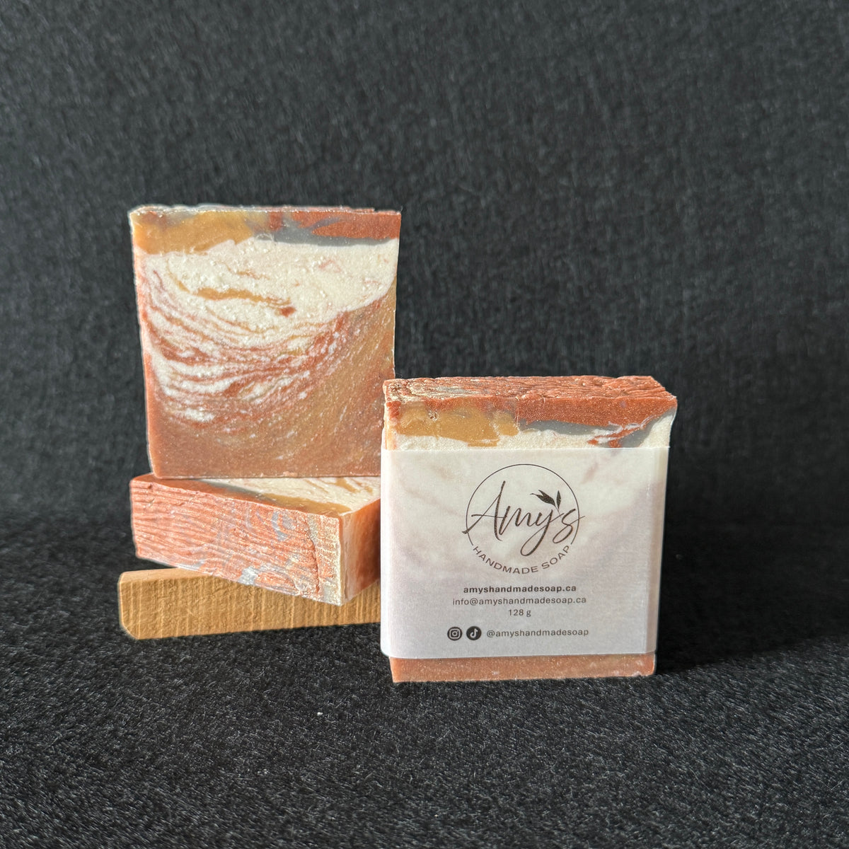 Three handmade soap bars with a terracotta and cream marbled pattern, displayed on a dark textured background. One bar is stacked on wooden blocks, and another has a paper label wrapped around it with the brand name 'Amy's Handmade Soap'.