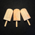 Three pale peach and cream popsicles with wooden sticks, arranged side by side on a dark textured background.