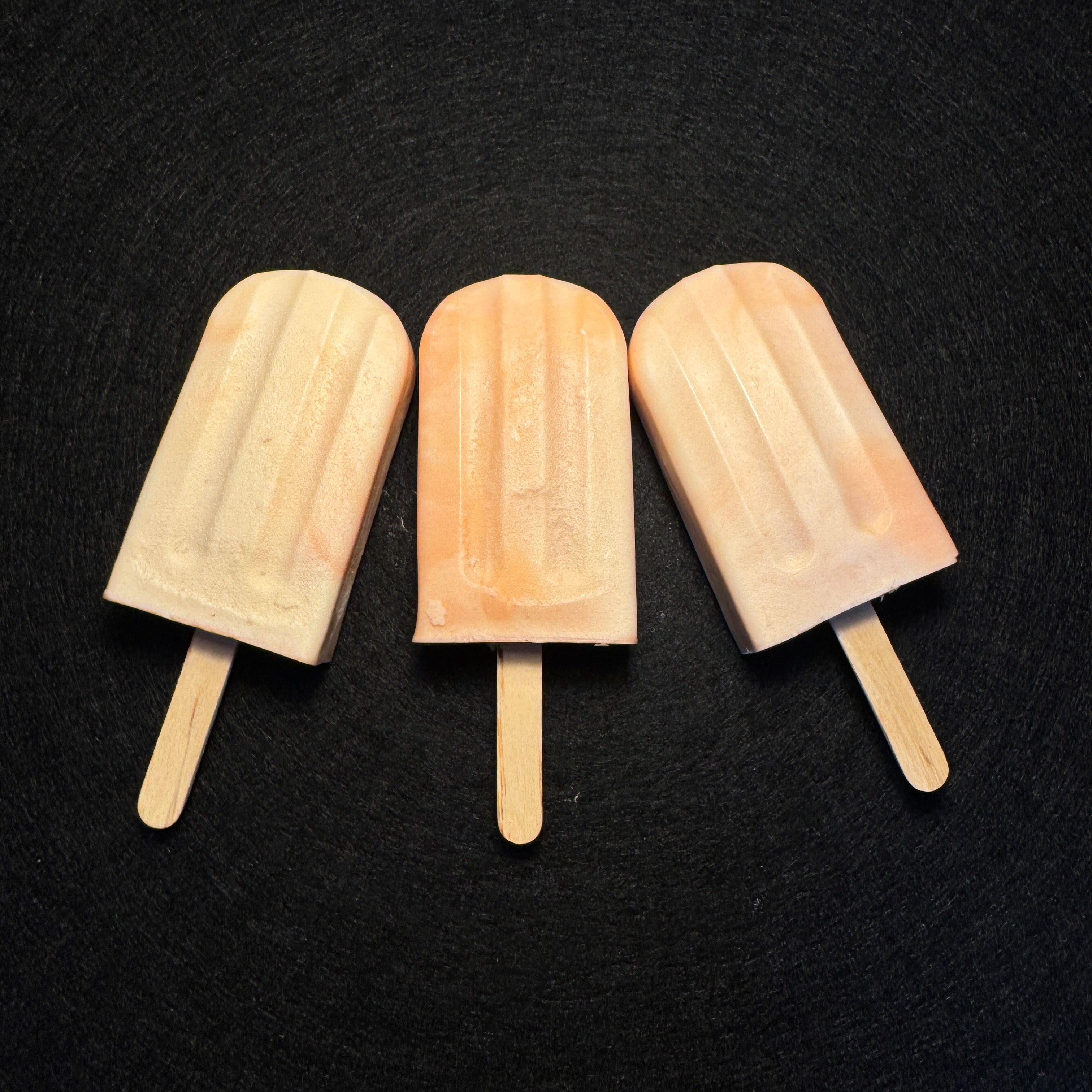Three pale peach and cream popsicles with wooden sticks, arranged side by side on a dark textured background.