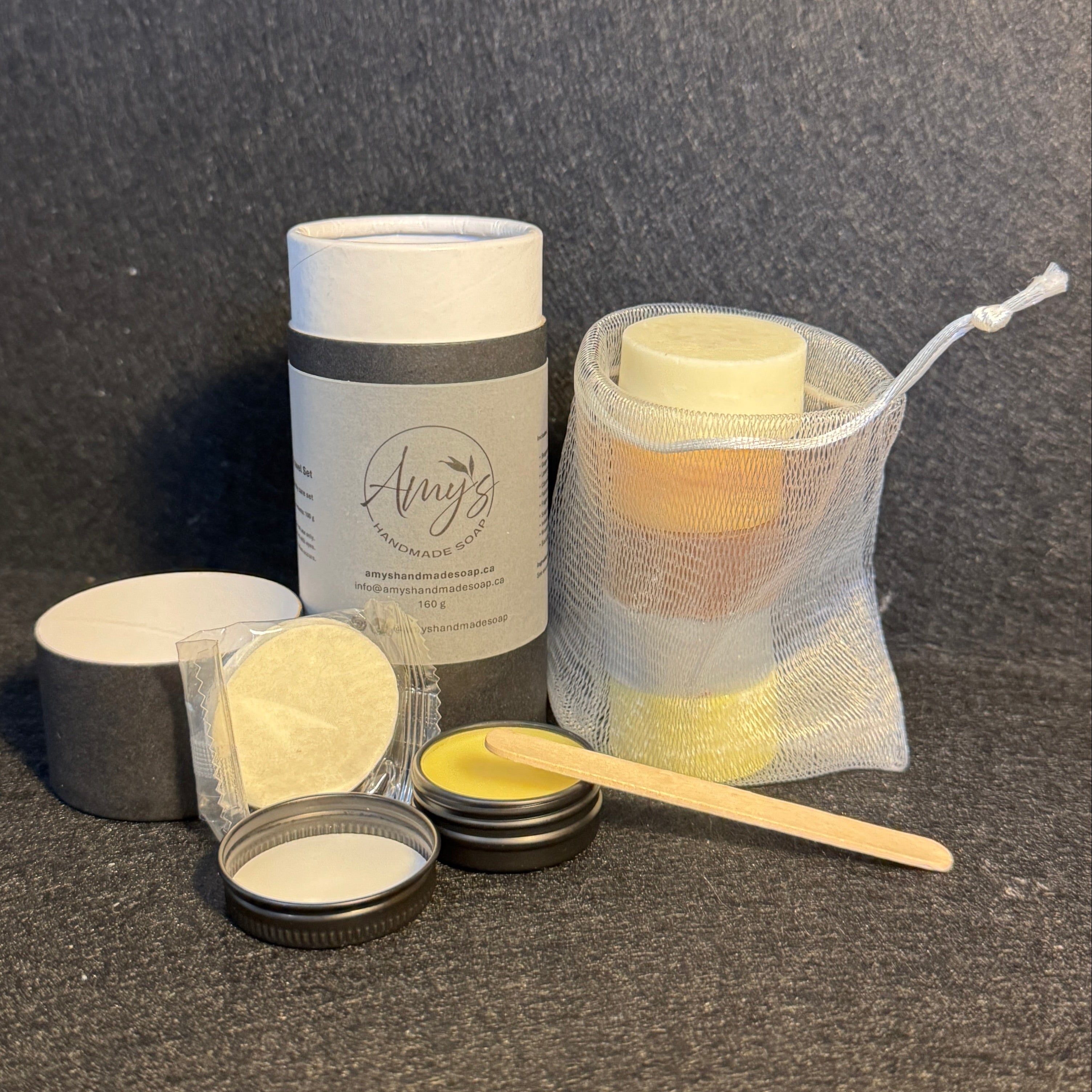 A set of skincare products arranged on a dark textured surface, including a cylindrical tube with a white lid, an open metal tin with yellow balm, and a mesh bag holding a selection of little soaps.